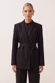 A woman modeling a black belted blazer with matching trousers, worn over a black and white patterned blouse, against a plain light beige background.