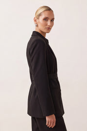 A woman modeling a black tailored blazer with a matching belt, shown in side profile against a plain light background.