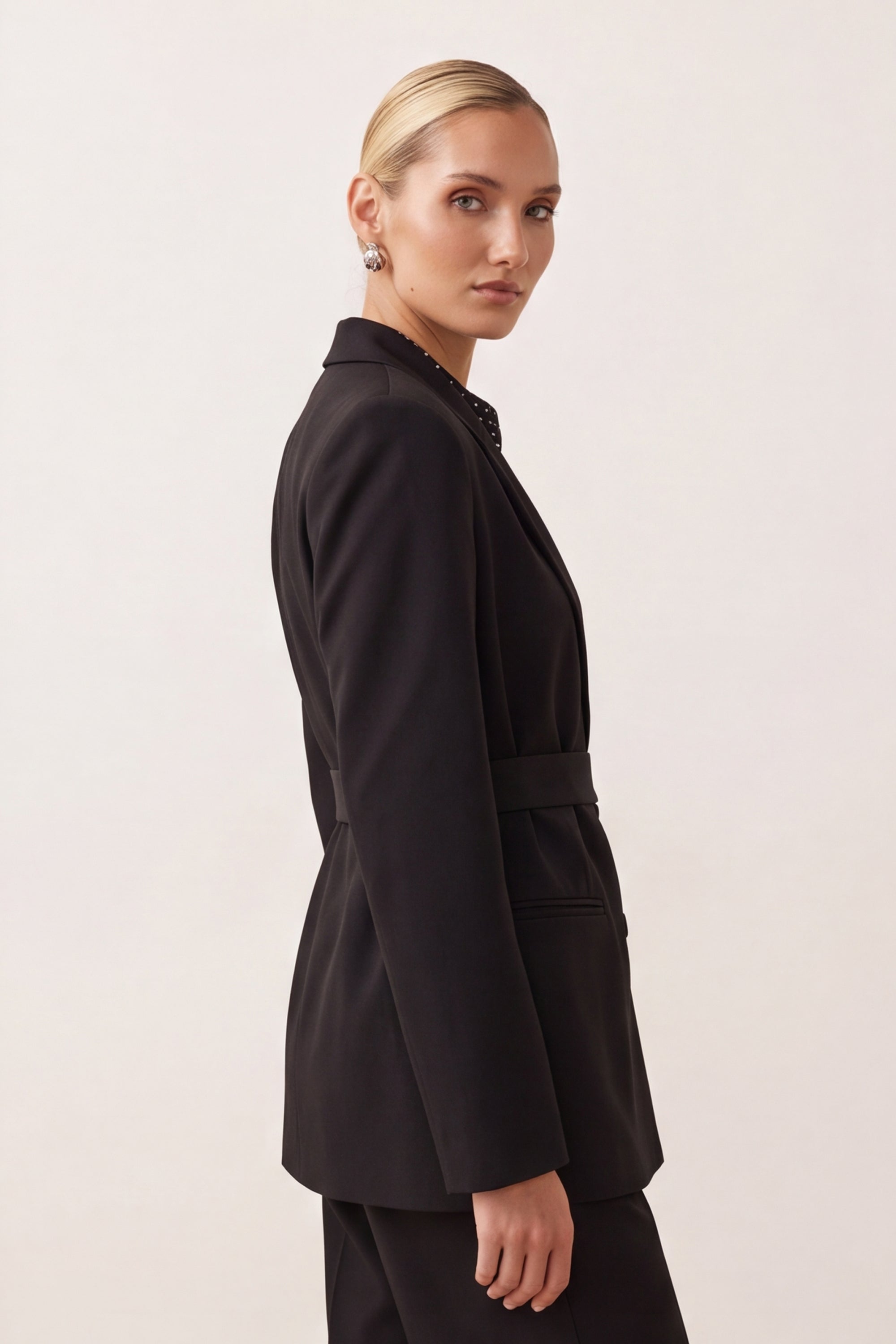A woman modeling a black tailored blazer with a matching belt, shown in side profile against a plain light background.