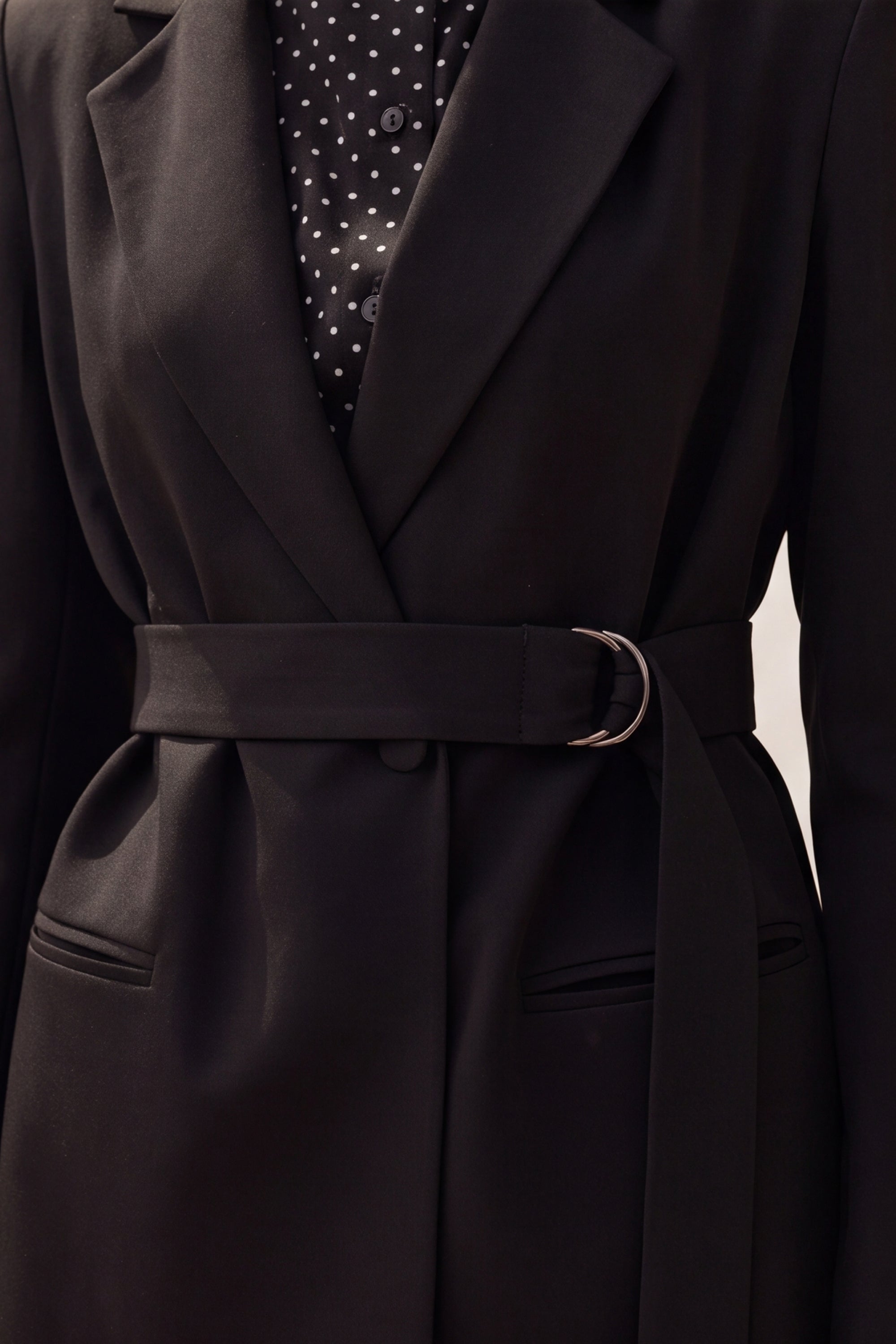 Close-up of a black belted coat with a silver-tone ring buckle, worn over a black and white polka dot shirt.