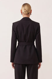 A woman shown from the back wearing a tailored black blazer with a matching belt and black trousers against a plain light background.