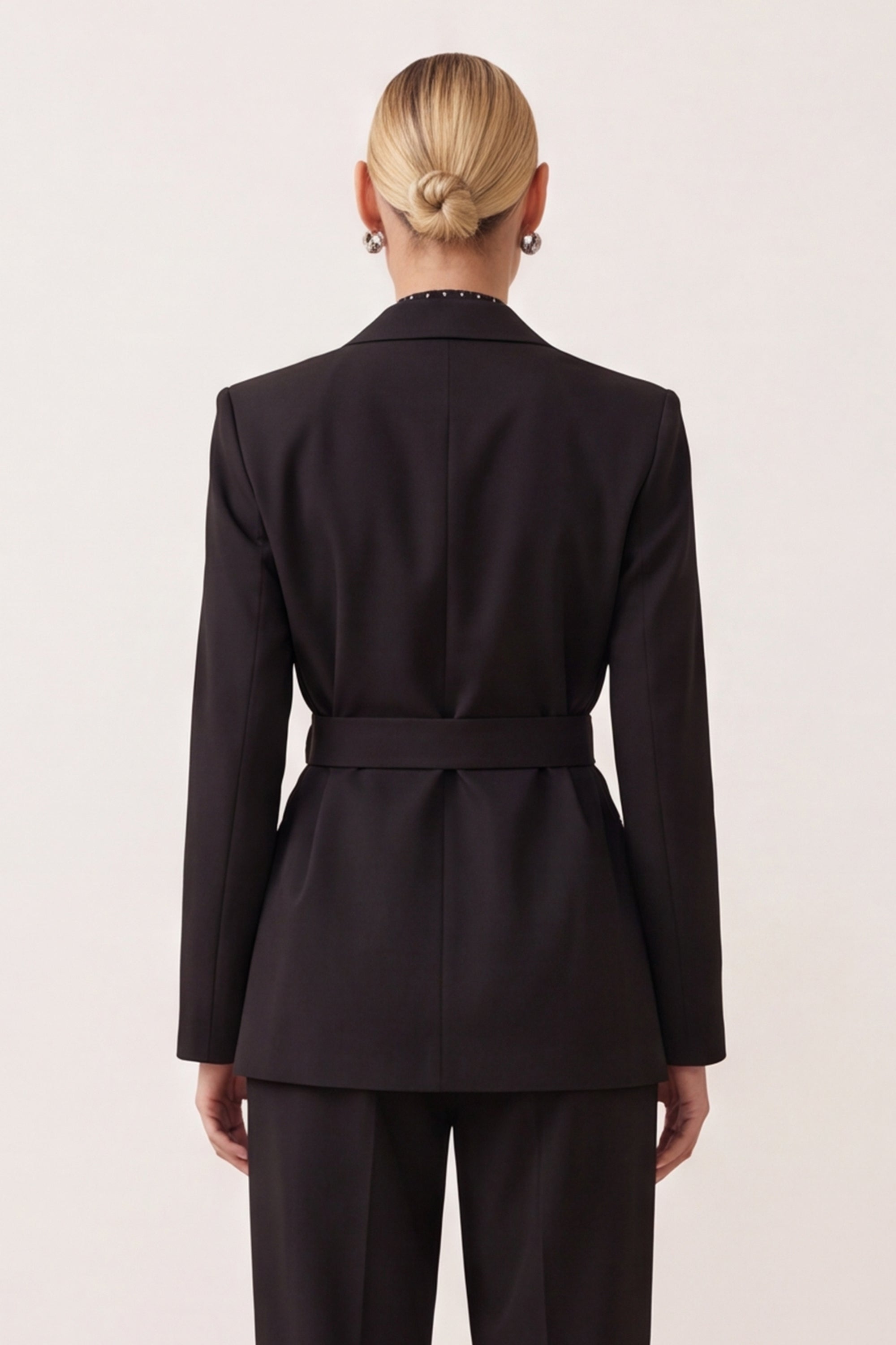 A woman shown from the back wearing a tailored black blazer with a matching belt and black trousers against a plain light background.