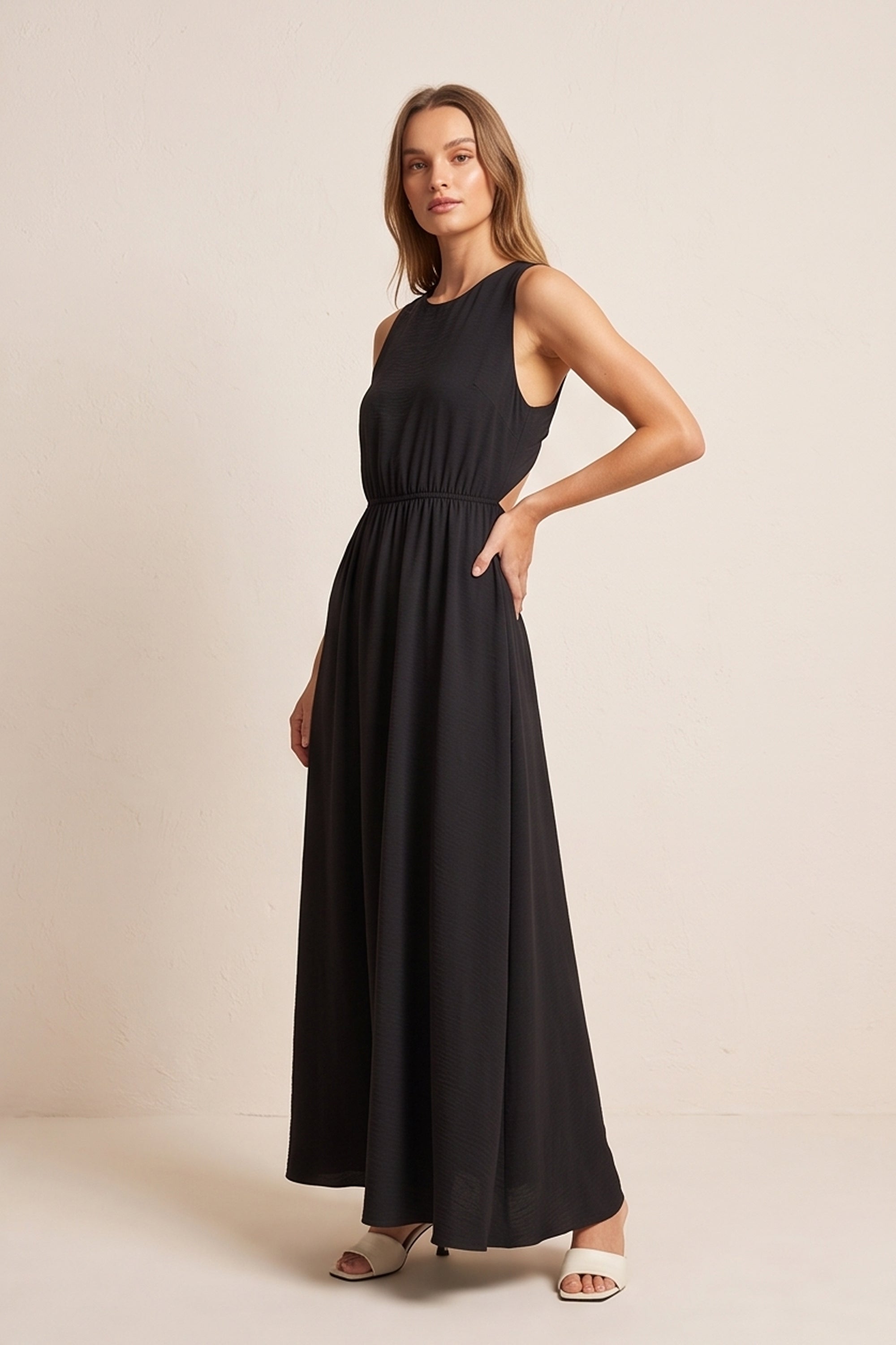 Side view of a woman wearing the black Virella Maxi Dress with sleeveless design, open back, and floor-length silhouette for elegant evening wear