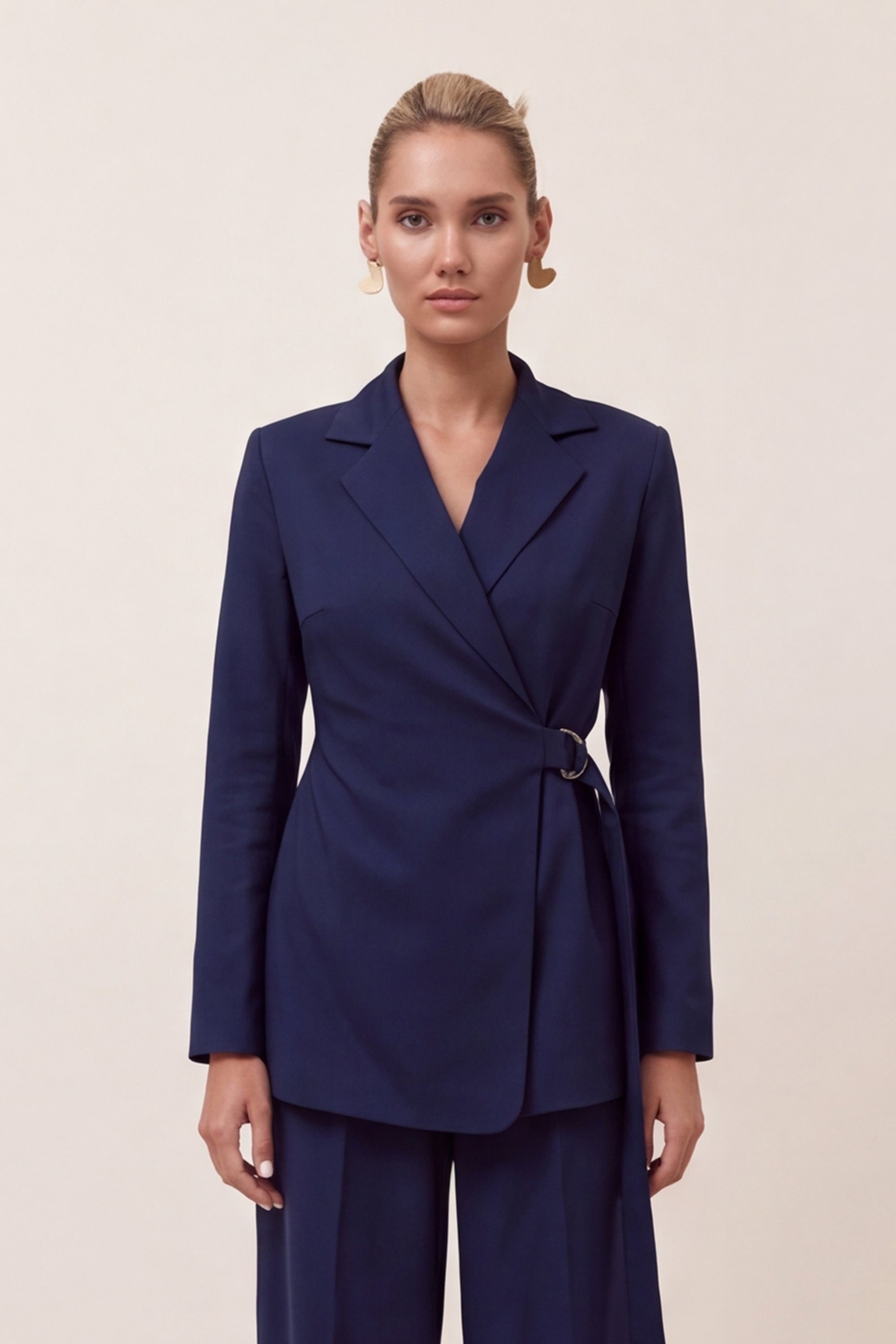 A woman modeling a navy blue wrap blazer with a matching pair of trousers, photographed against a plain beige studio background.