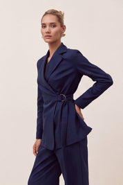 A woman modeling a navy blue tailored suit set consisting of a belted wrap-style blazer and matching trousers, posed against a plain light beige studio background.