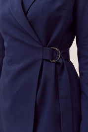Close-up of a navy blue wrap-style dress or tunic with a matching waist belt and a prominent circular metallic ring detail at the center, shown on a model against a light neutral background.