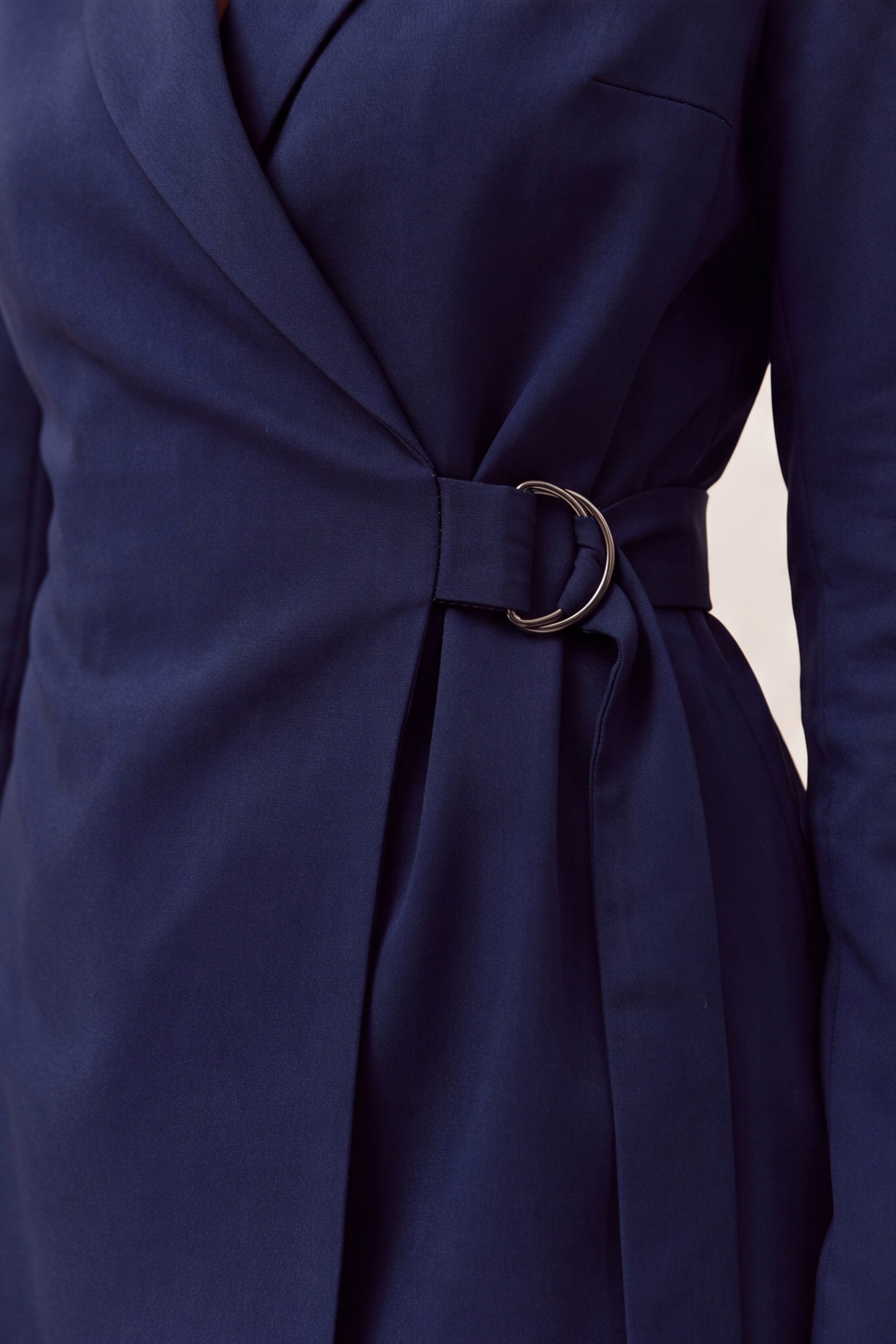 Close-up of a navy blue wrap-style dress or tunic with a matching waist belt and a prominent circular metallic ring detail at the center, shown on a model against a light neutral background.