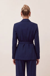 A woman viewed from the back wearing a navy blue tailored blazer with a matching waist tie and matching trousers, against a plain light beige studio background.