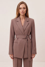 A woman modeling a taupe-brown belted blazer and matching trousers against a plain light beige studio background.