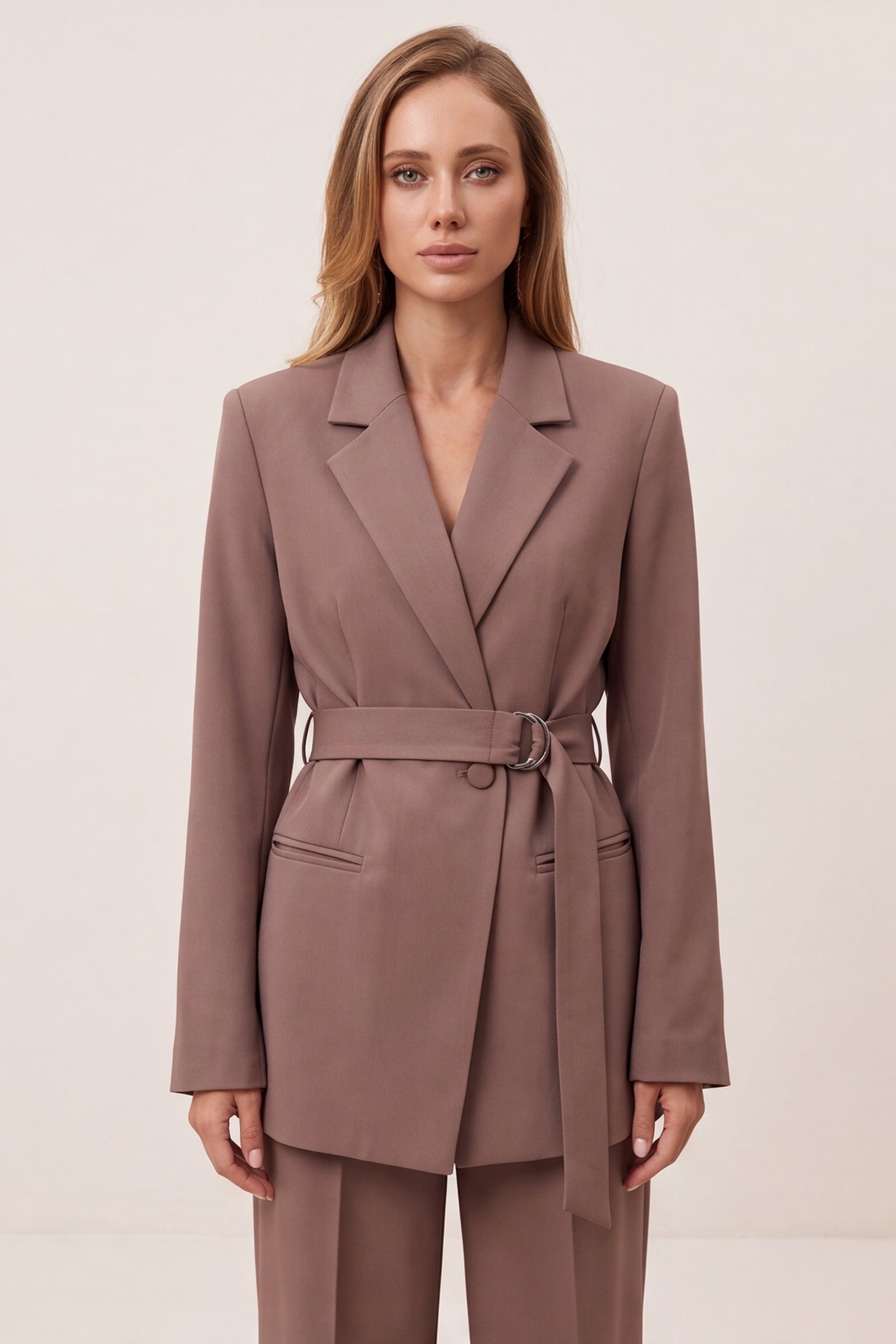 A woman modeling a taupe-brown belted blazer and matching trousers against a plain light beige studio background.