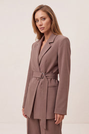 A woman modeling a taupe-brown belted blazer with matching trousers against a plain, light beige studio background.