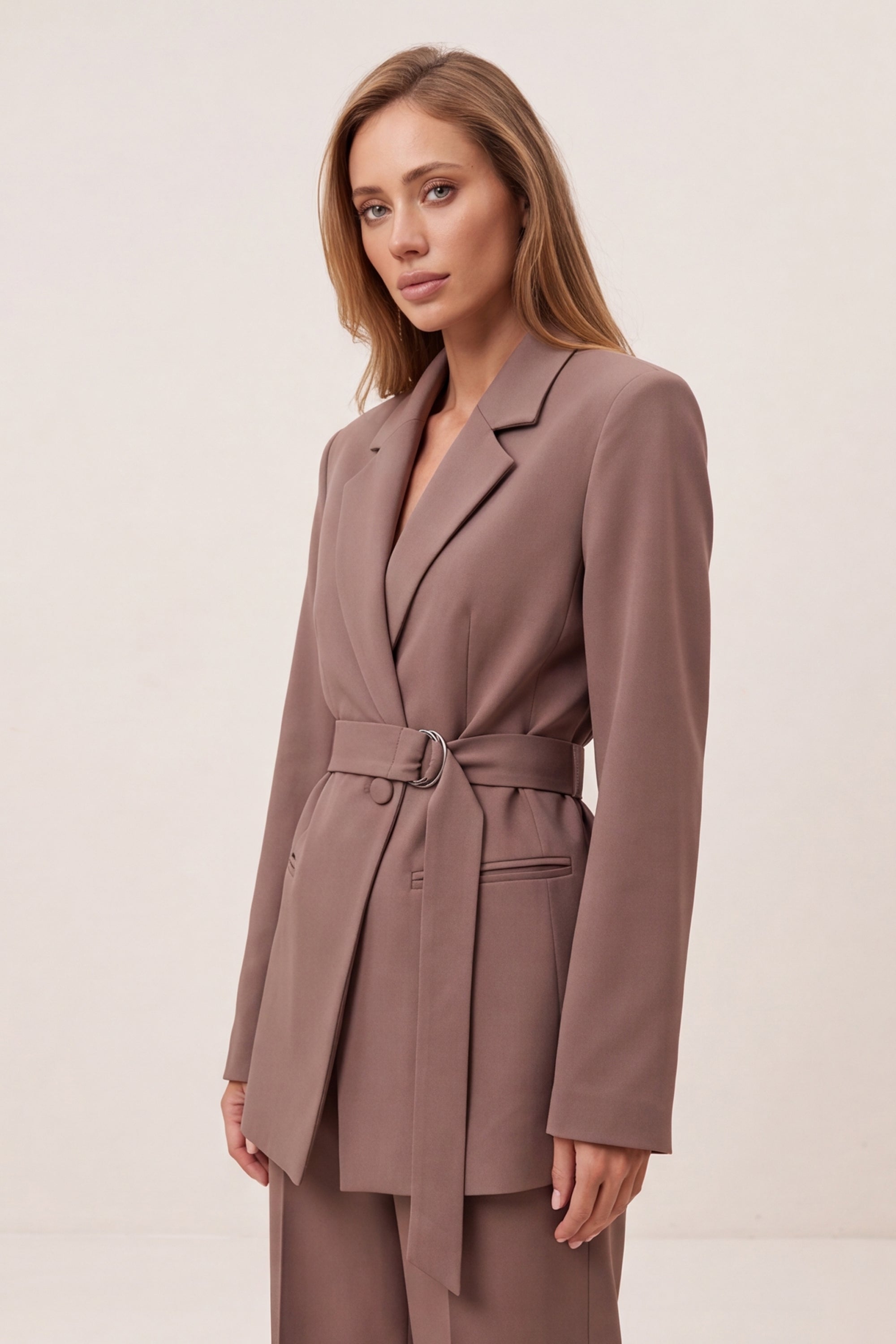 A woman modeling a taupe-brown belted blazer with matching trousers against a plain, light beige studio background.