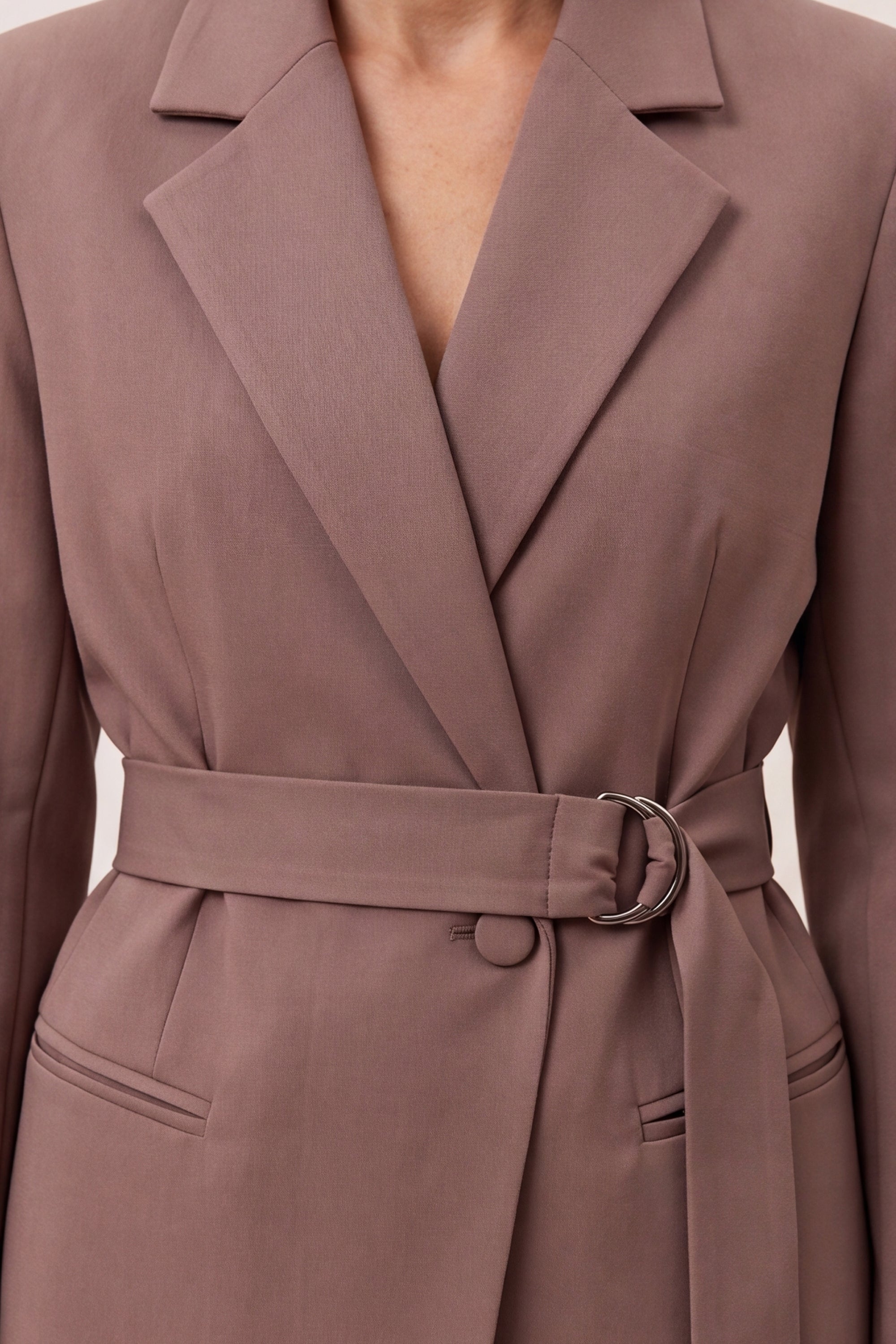 Close-up of a taupe-brown belted wrap coat with notched lapels and a silver-tone ring detail at the waist, shown on a model.