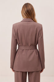 A woman shown from the back wearing a tailored blazer and matching trousers in a muted dusty mauve-brown color, against a plain light beige studio background.