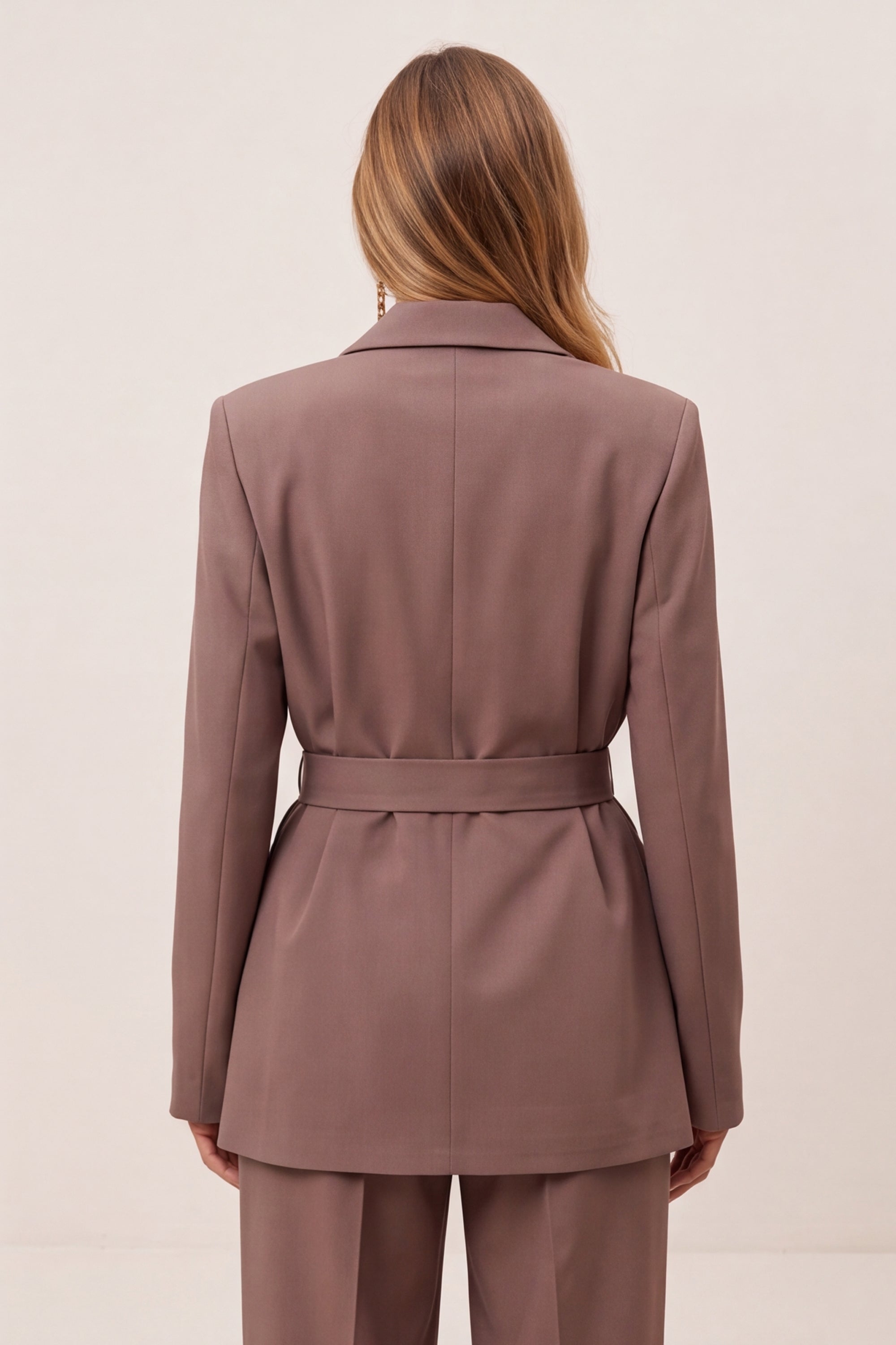 A woman shown from the back wearing a tailored blazer and matching trousers in a muted dusty mauve-brown color, against a plain light beige studio background.