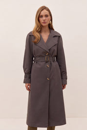 A woman modeling a dark gray belted trench coat with a double-breasted front and wide lapels, photographed against a plain light beige studio background.
