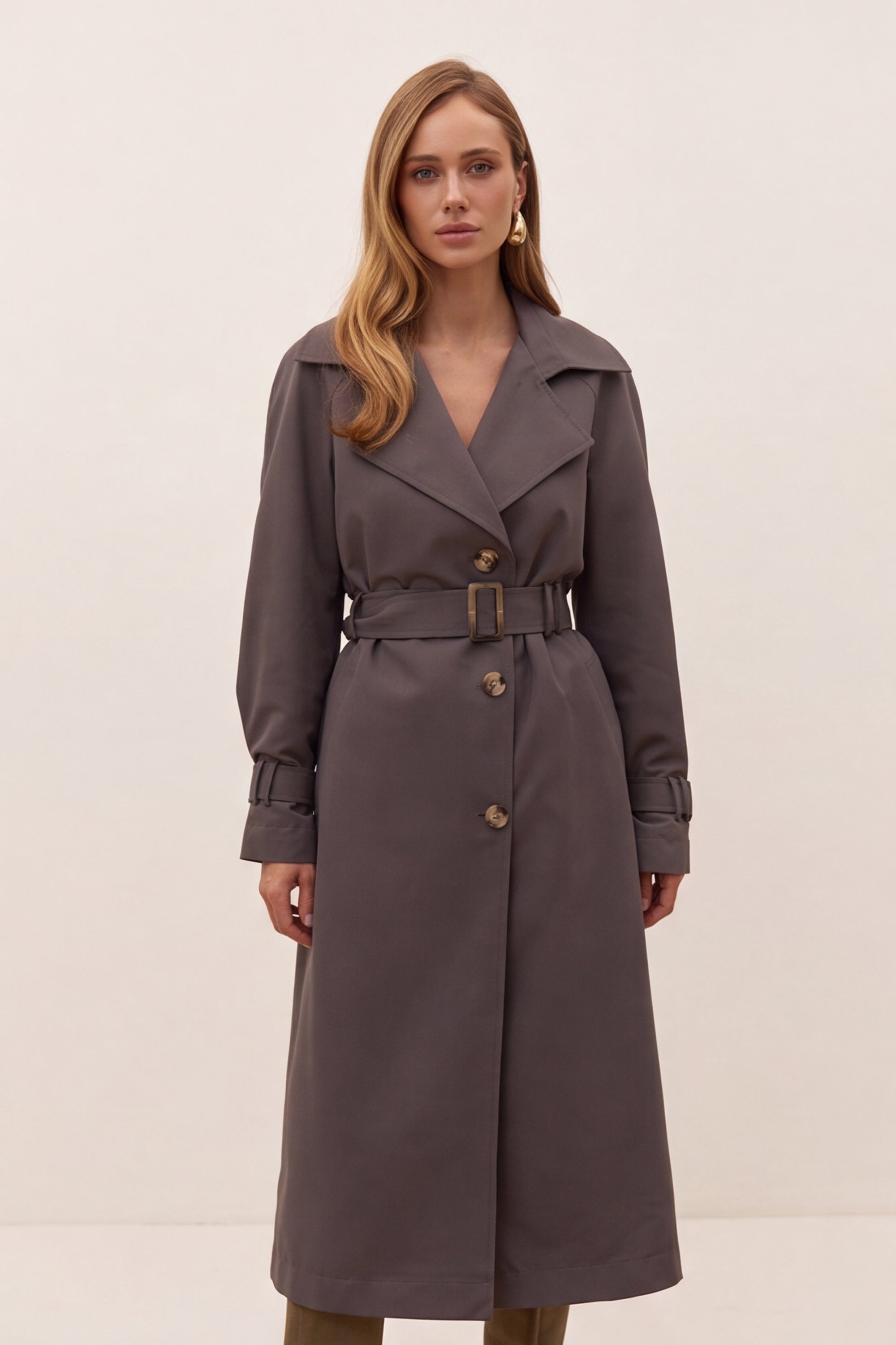 A woman modeling a dark gray belted trench coat with a double-breasted front and wide lapels, photographed against a plain light beige studio background.