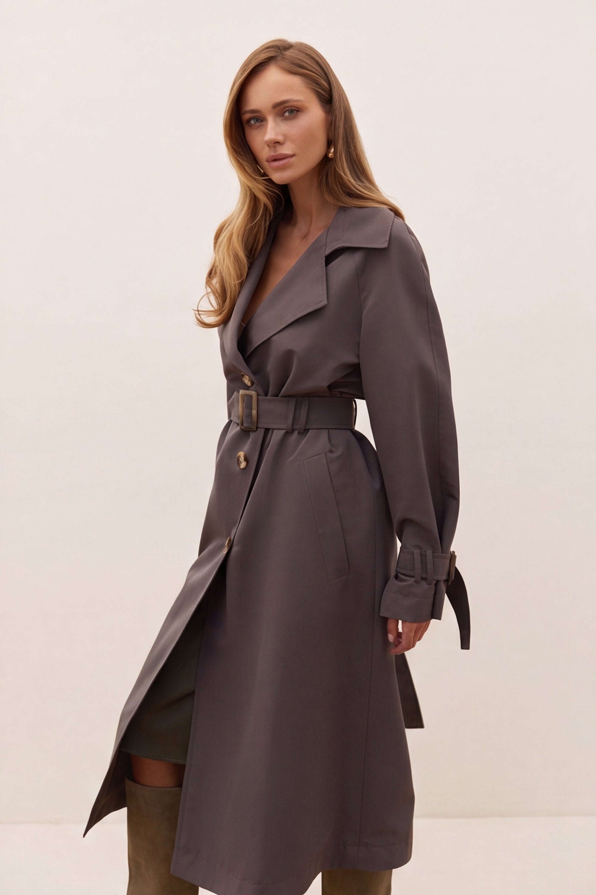 A woman modeling a dark gray belted trench coat with wide lapels and a wrap front, photographed against a plain light beige studio background.