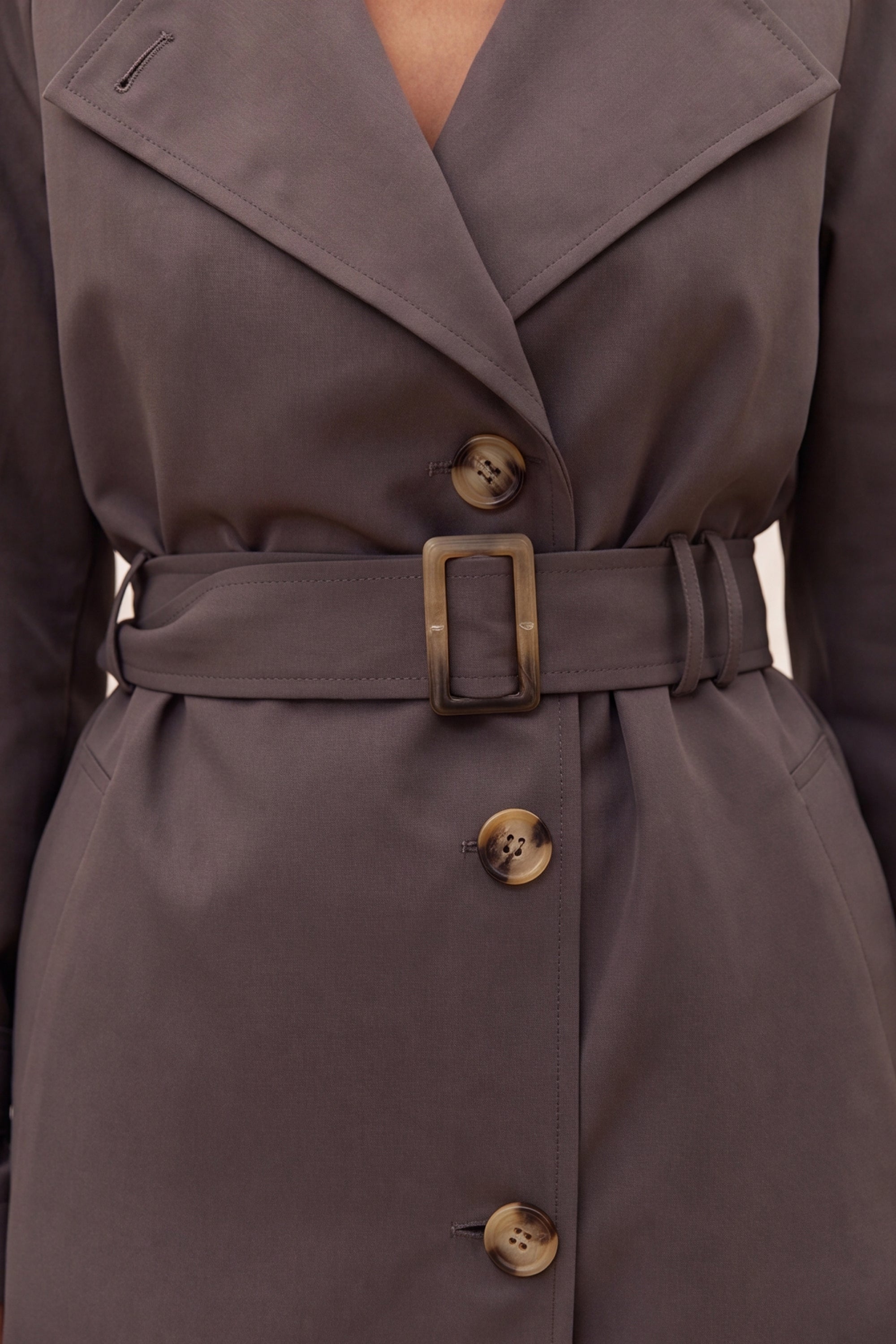 Close-up of a dark gray belted trench coat with large bronze-toned buttons and a matching belt buckle, shown on a model.