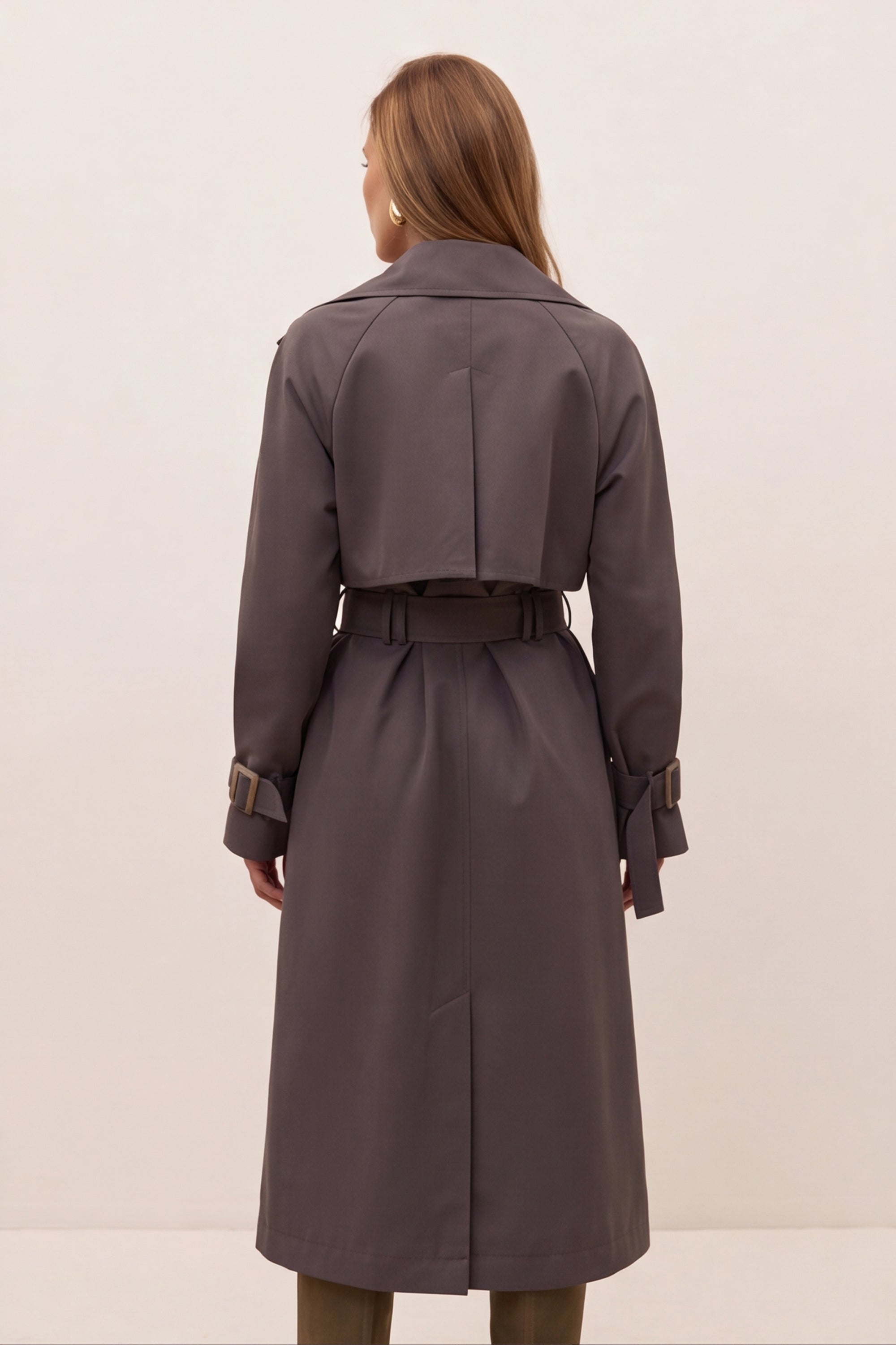 Back view of a dark gray belted trench coat with large bronze-toned buttons and a matching belt buckle, shown on a model.