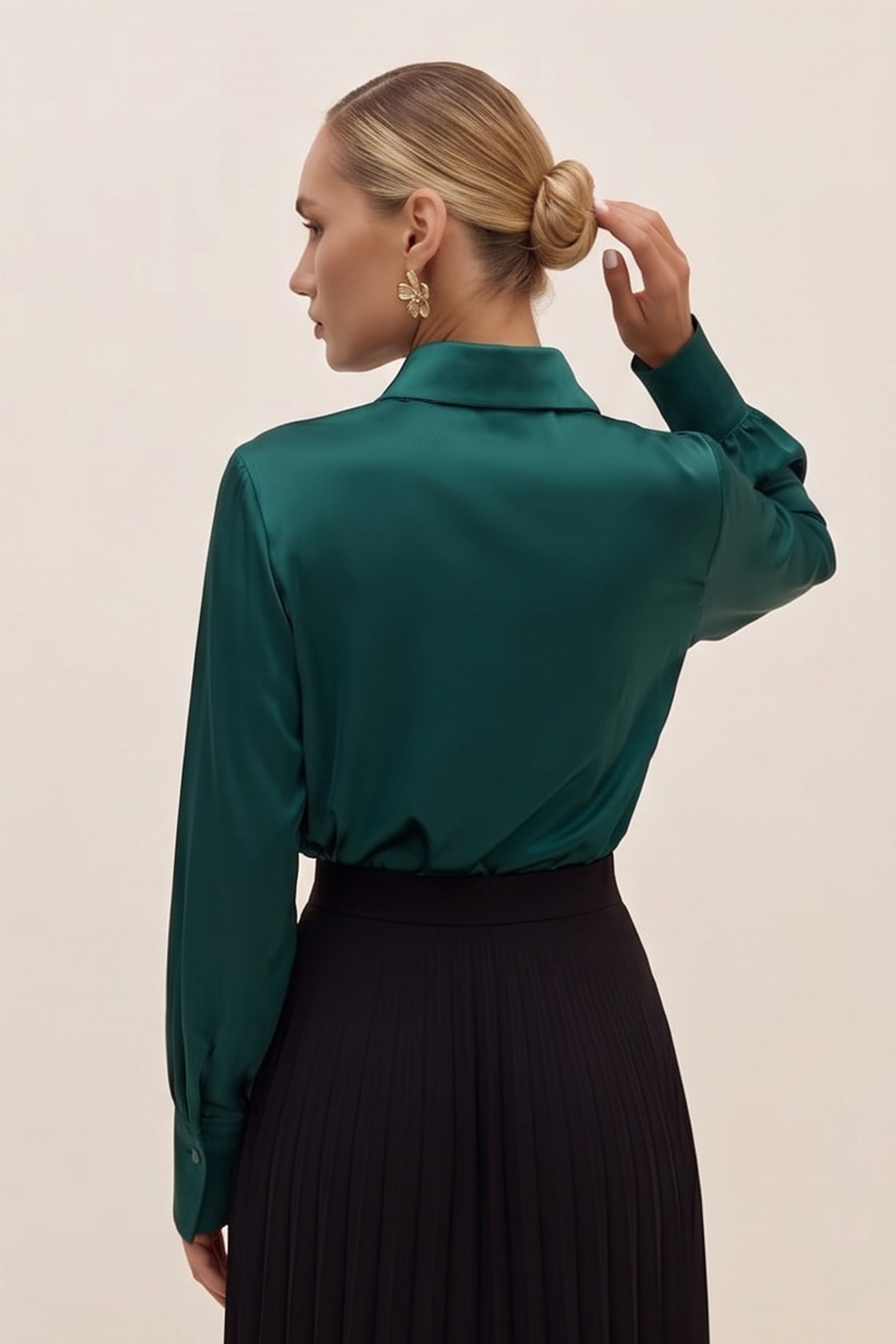 Back view of a woman wearing a silk like green eminence blouse by femeador and black skirt against a beige background