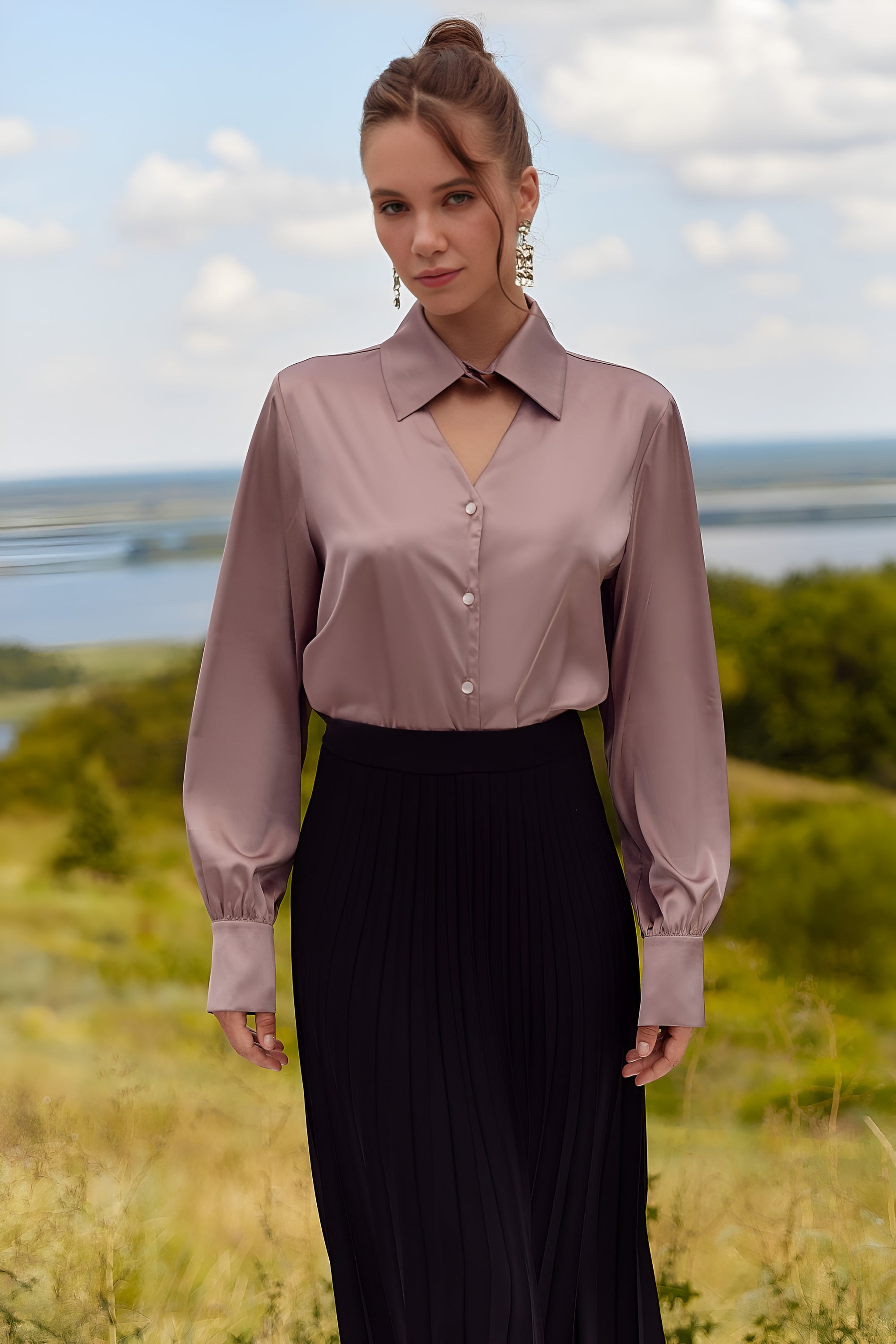 Front view of a woman wearing a light pink blouse and black pants standing in a scenic outdoor setting.