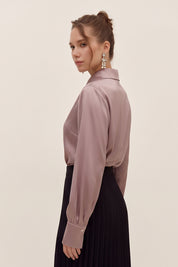 Side view of a woman wearing a light silk like pink eminence blouse by femeador and black skirt against a beige background