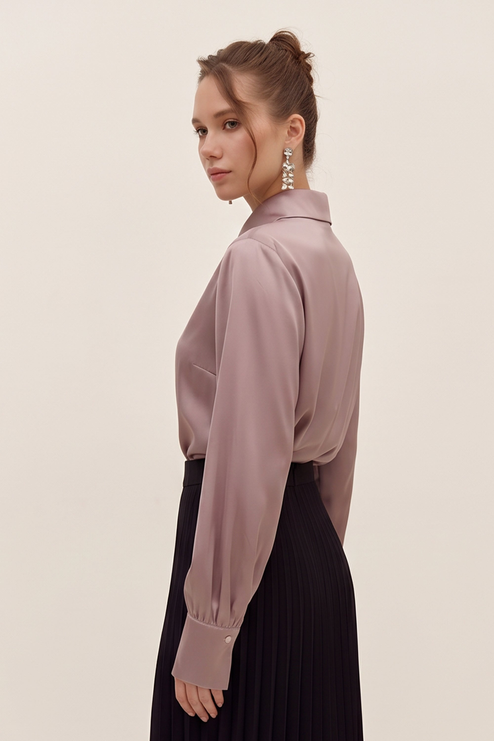 Side view of a woman wearing a light silk like pink eminence blouse by femeador and black skirt against a beige background
