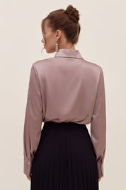 Back view of a woman wearing a light silk like pink eminence blouse by femeador and black skirt against a beige background