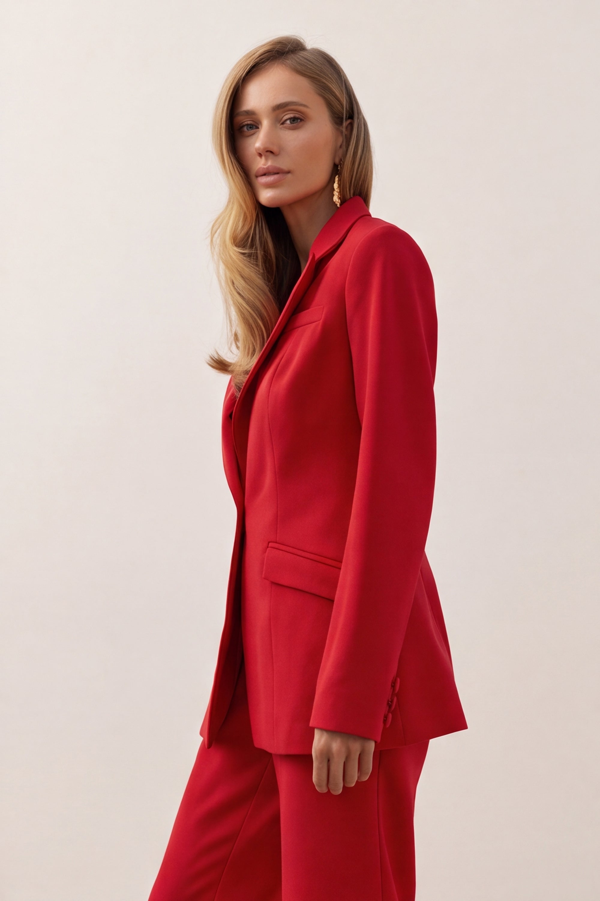 A woman shown from the side wearing a bright red tailored blazer and matching red trousers against a plain light beige studio background.