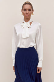 Front view of a woman wearing a white reverie blouse by femeador with a bow tie and a blue pleated skirt on a beige background
