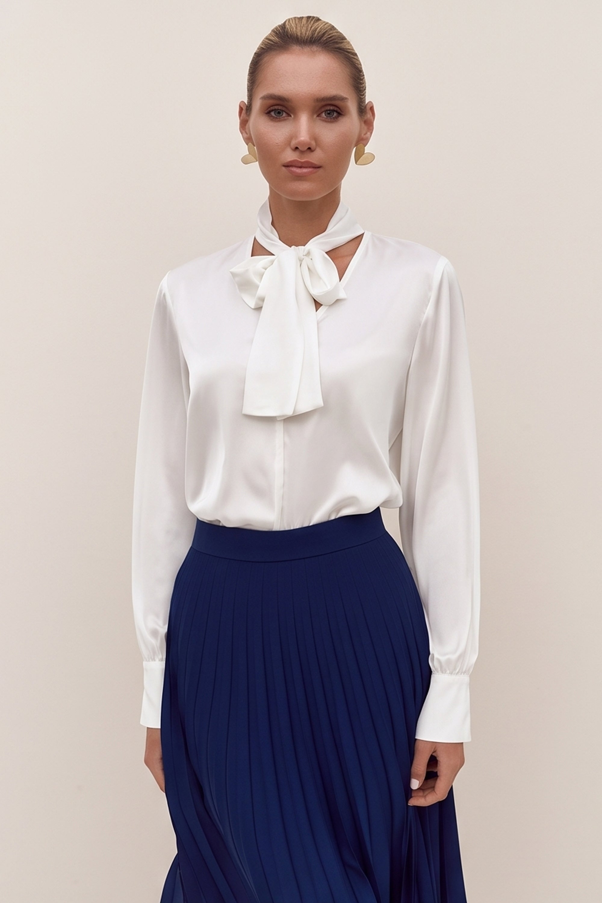 Front view of a woman wearing a white reverie blouse by femeador with a bow tie and a blue pleated skirt on a beige background