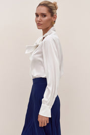 Side view of a woman wearing a white reverie blouse by femeador with a bow tie and a blue pleated skirt on a beige background