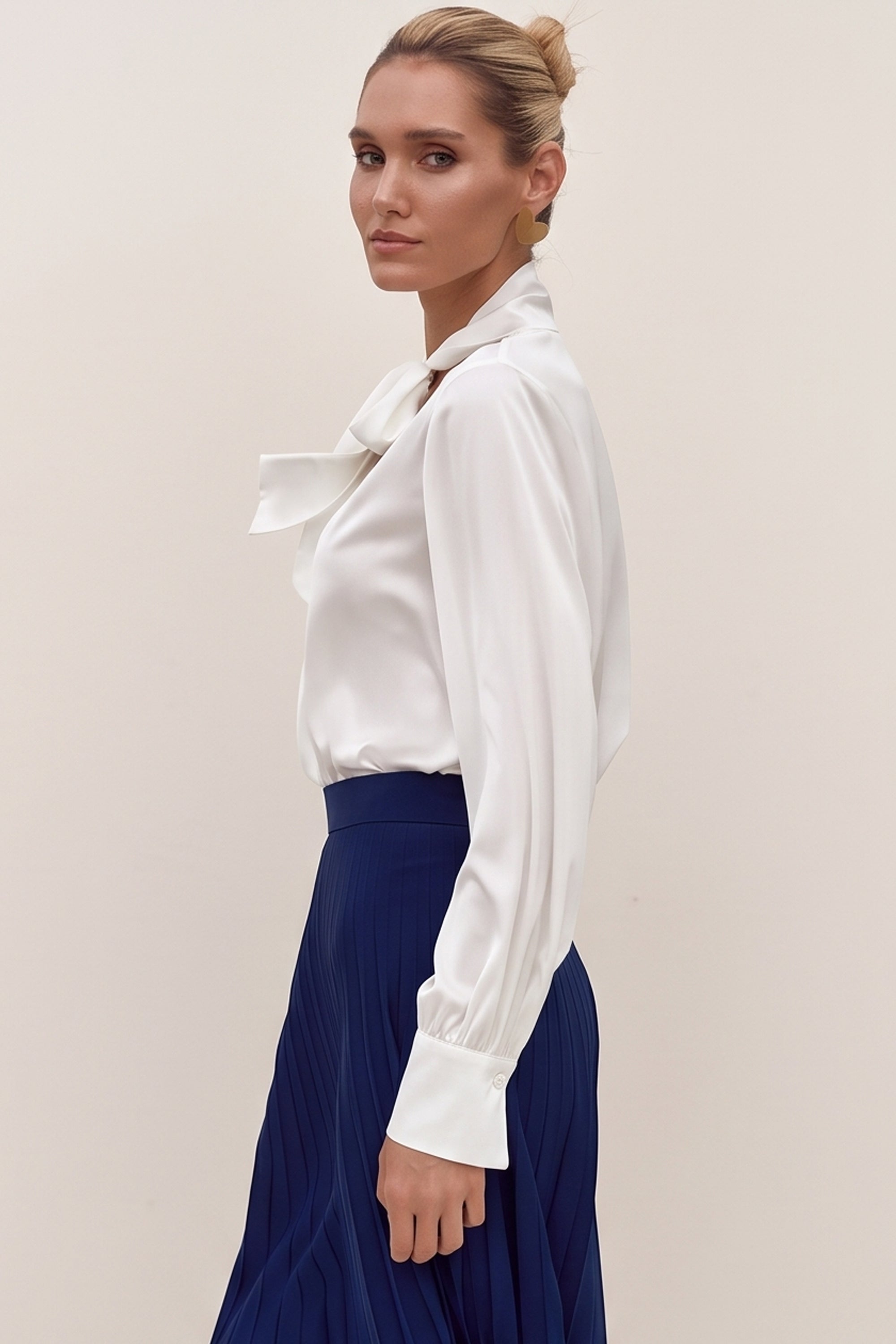 Side view of a woman wearing a white reverie blouse by femeador with a bow tie and a blue pleated skirt on a beige background