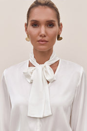 Close up view of a woman wearing a white reverie blouse by femeador with a bow tie and a blue pleated skirt on a beige background