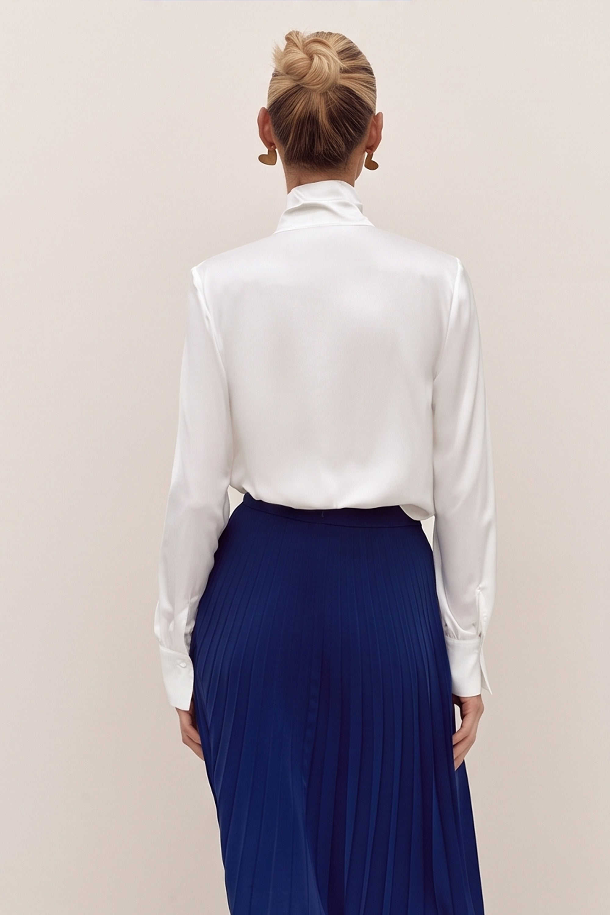 Back view of a woman wearing a white reverie blouse by femeador with a bow tie and a blue pleated skirt on a beige background