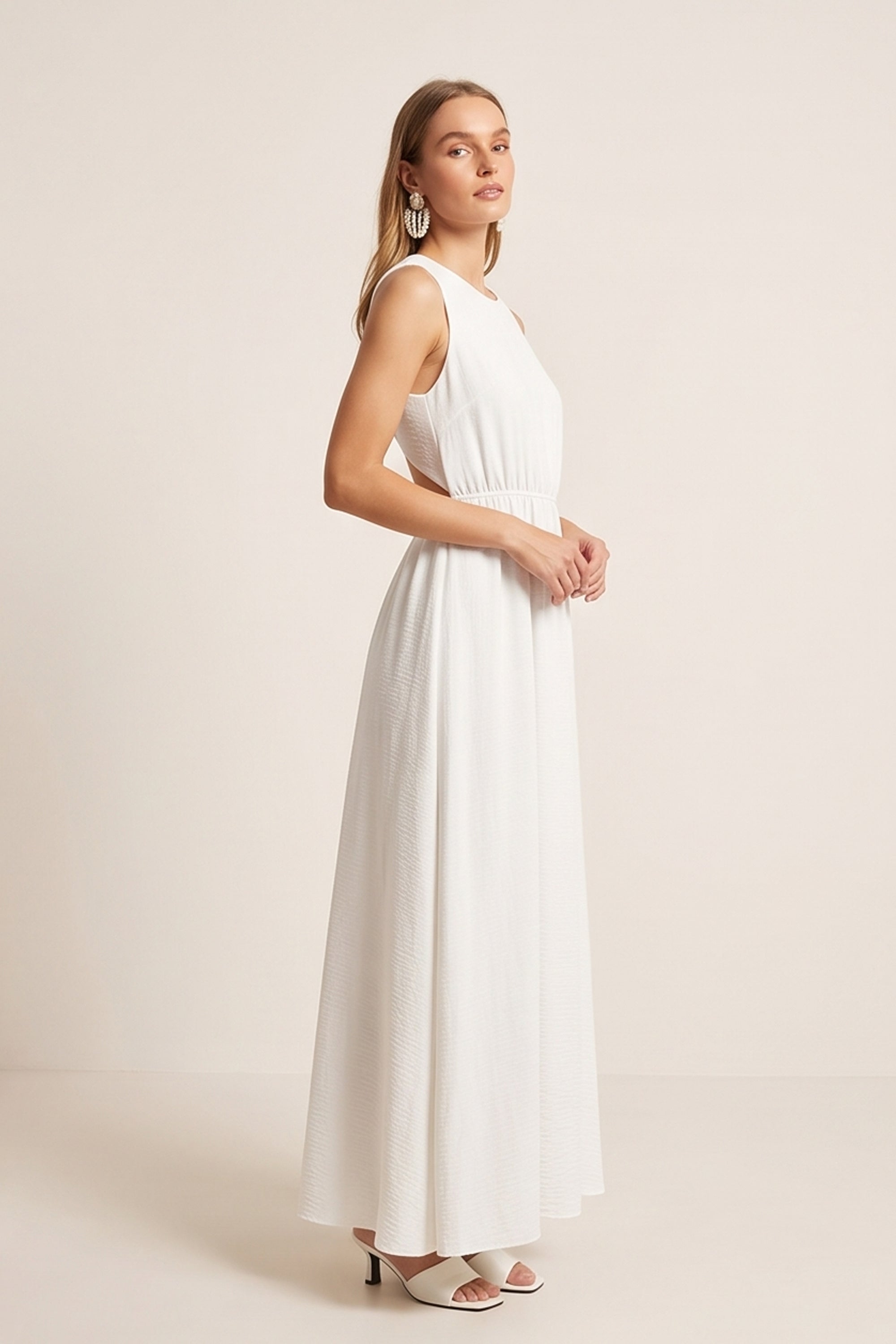 Side view of a woman wearing the white Virella Maxi Dress with sleeveless design, open back, and floor-length silhouette for elegant evening wear